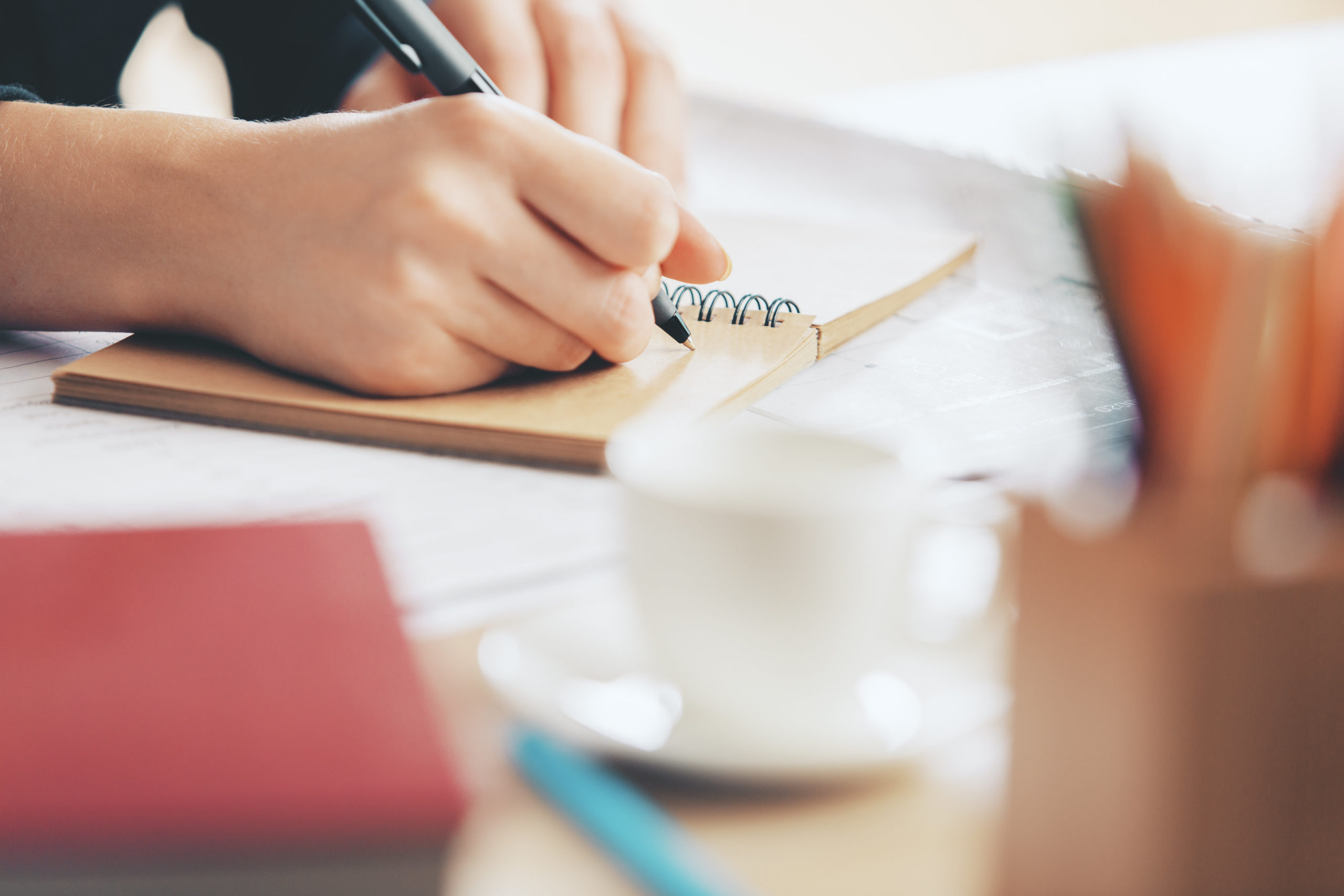 Close up of female's hands writing scholarship letter of recommendation in spiral notepad placed on wooden desktop with coffee cup and other items